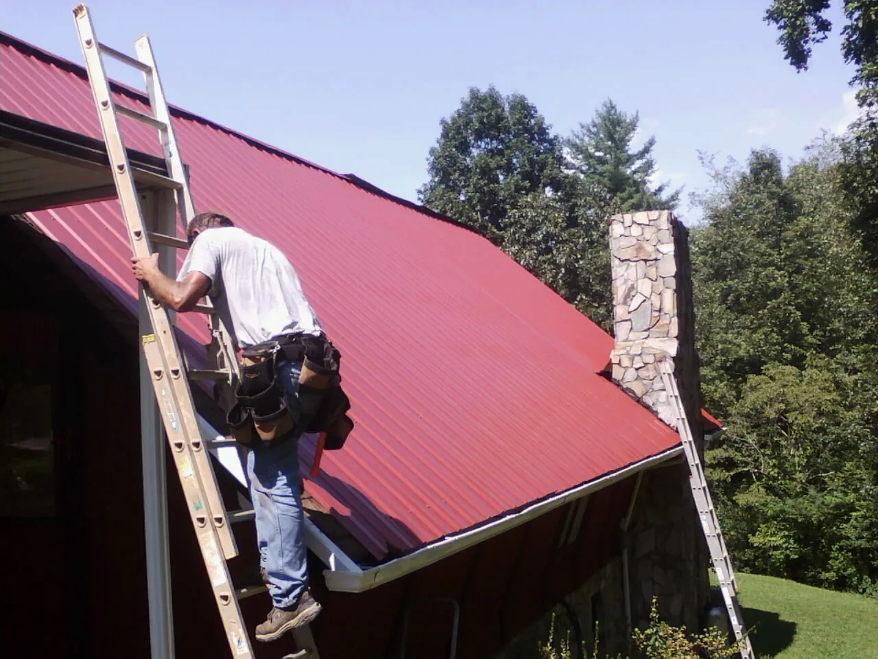 Roofer on a ladder beside a red metal roof and stone chimney — Soffit & Fascia Repair by Roofing Brothers in Bonaire